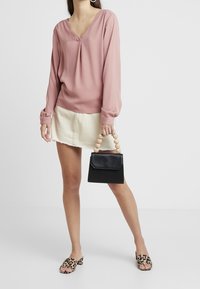 Blusa lunga a maniche con scollatura a V di colore rosa tenue, abbinata a una gonna di denim chiaro. Tiene una borsa nera con manico in perline di legno e indossa sandali con stampa leopardata.