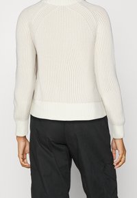 Cremfarbener gerippter Pullover mit hohem Kragen, enganliegenden Ärmelbündchen und einem Taillenbund. Die Textur ist weich und zeigt vertikale Streifen.