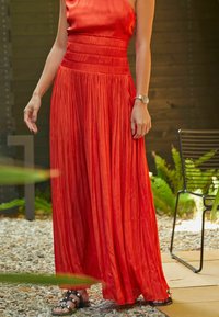 Robe maxi plissée rouge avec un corsage ajusté et une ceinture à la taille. Tissu en satin avec une texture lisse. Portée avec des sandales noires et une montre.