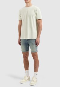 Hellgrünes T-Shirt mit kurzen Ärmeln, kombiniert mit blauen Denim-Shorts in verwittertem Design. Legere weiße Sneakers mit hellen Akzenten.