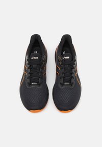 Crne atletske tenisice s teksturiranim mrežastim gornjim dijelom, narančastim dodacima i zatvaranjem na vezicu. Uključuju Gore-Tex detalje za vodonepropusnost.