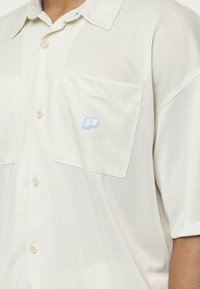 Koszula na krótki rękaw na guziki w kolorze jasnego kremu, z tkaniny o fakturze. Posiada dwie naszywane kieszenie i jasnoniebieskie logo "P".