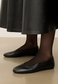 Ballerines en cuir noir avec un bout arrondi, texture lisse et profil bas, portées avec des collants noirs transparents et une longue jupe noire.