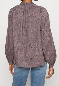 Blouse fluide avec un col smocké, des manches bouffantes longues et un motif géométrique dans des tons atténués de rose et de marine sur un tissu léger.