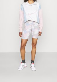 Sweat-shirt à zip quart Nike en blanc, rose et bleu ; associé à un short de vélo camouflé pastel. Baskets blanches avec des accents gris et des détails rouges.
