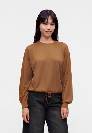 Jeune femme aux longs cheveux foncés portant un pull marron avec cordon de serrage à la taille et un jean noir, debout devant un fond clair uni.