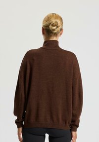 Kobieta z blond włosami w kok, nosząca złote kolczyki w kształcie kół i luźny brązowy sweter z golfem, zwrócona tyłem na jednolitym tle.