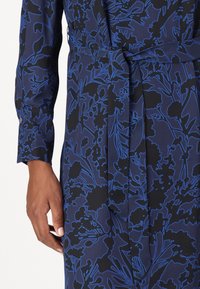 Mörkblå klänning med ett blommönster i ljusare blå nyanser. Tyget verkar slätt och har en knytning i midjan. Långa ärmar med knappmanchetter.