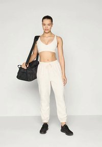 Ung kvinde i hvid sports-bh og joggers holder en sort træningstaske over skulderen, iført sorte sportsko, står foran en ensfarvet baggrund.