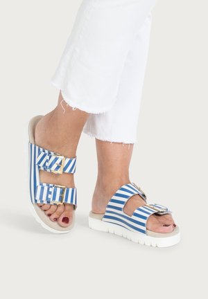 Feet wearing blue and white striped slide sandals with gold buckles, paired with white cropped frayed hem pants.