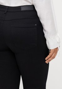 Black stretchy jeans with a back pocket, smooth texture, and a leather waistband label. A white long-sleeve shirt is partially visible.