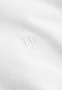 Tela blanca con una superficie texturizada. Incluye una pequeña 'U' bordada con costura sutil. La vista en primer plano resalta el detalle del material.