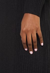 Schwarzes, gerippte Strickbekleidung mit einer strukturierten Oberfläche, daneben eine Hand mit hellroten Nägeln, die neben dem Stoff positioniert ist.