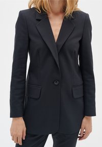 Černý přizpůsobený blazer s výstřihem do V, jednobuttonovým zapínáním, dvěma předními kapsami a štíhlým střihem, vyrobený z hladkého materiálu.