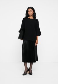 Pull noir surdimensionné avec des manches larges, associé à une jupe midi plissée noire. Mannequin tenant un sac à main noir et portant des talons noirs.