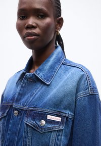 Γυναίκα με πλεξούδες φοράει ένα μπλε τζιν μπουφάν Calvin Klein με κουμπωτές τσέπες στο στήθος και χρυσούς κρίκους στα αυτιά, σε απλό φόντο.