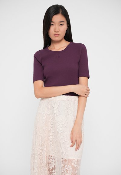 Top violet cu mânecă scurtă, ajustat, cu textură canelată și un logo subtil embossat; asociat cu o fustă din dantelă florală transparentă în nuanțe de roz deschis.