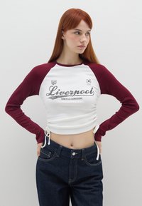 Witte en bordeauxrode langemouwen shirt met "Liverpool" grafiek, gerimpelde zijkanten, gecombineerd met donkerdenim hoge taille jeans.