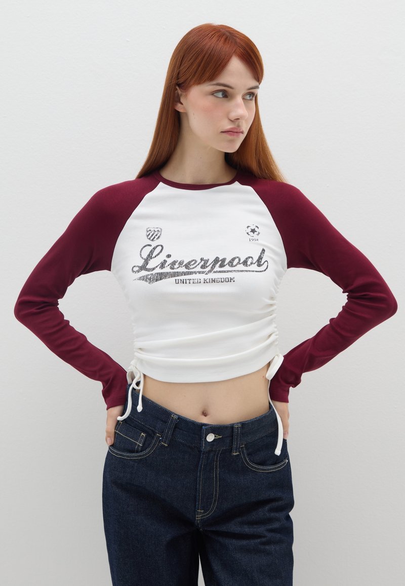 Witte en bordeauxrode langemouwen shirt met "Liverpool" grafiek, gerimpelde zijkanten, gecombineerd met donkerdenim hoge taille jeans.