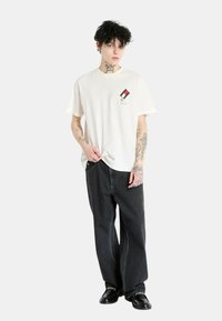 Camiseta de algodón blanca con un pequeño diseño bordado en el lado izquierdo del pecho, combinada con pantalones de mezclilla negros holgados y zapatos negros.