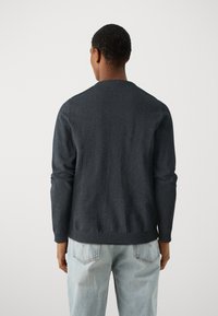 Maglione grigio scuro con scollatura rotonda, maniche lunghe e orlo a coste. Indossato sopra jeans di denim azzurro chiaro. Design minimalista.