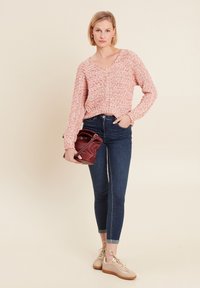 Bluse mit floralem Muster in Pink und V-Ausschnitt, langen Ärmeln, kombiniert mit dunkelblauen Skinny-Jeans und beigen Sneakers, hält eine rote Handtasche.