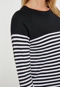 Schwarz-weißer gestreifter Pullover mit rundem Ausschnitt und geripptem Bündchen, der abwechselnd dicke und dünne Streifen aufweist. Weiche Stricktextur.