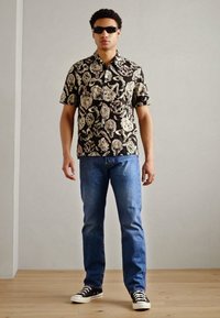 Schwarz bedrucktes Kurzarmhemd mit weißen und grünen Mustern, kombiniert mit blauen Jeans und schwarz-weißen Sneakers. Sonnenbrille getragen.