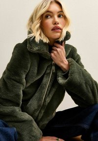 Mujer rubia con una chaqueta verde oscura y peluda de cuello alto, sosteniendo el cuello cerca de su rostro, sentada con vaqueros oscuros.