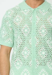 Chemise en crochet verte menthe à manches courtes avec un motif floral, fermeture à boutons sur le devant et un col texturé. Légère et en tissage ouvert.