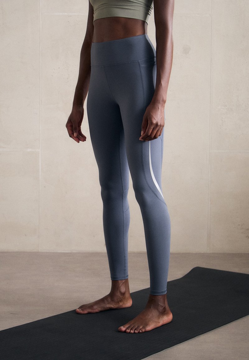 Leggings ajustés taille haute en tissu gris clair mat avec une rayure latérale blanche. Surface texturée, conçue pour le sport.