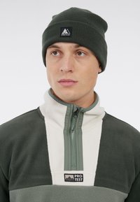 Groene beanie en een fleece jack met rits met witte en donkergroene panelen. Fleece textuur met een logo patch op de borst en rits.