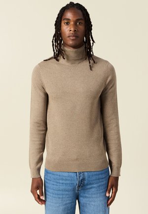 Beige Rollkragenpullover aus glattem Strickstoff. Figurbetonter Schnitt mit langen Ärmeln sowie gerippten Bündchen und Saum. Kombiniert mit Jeans.