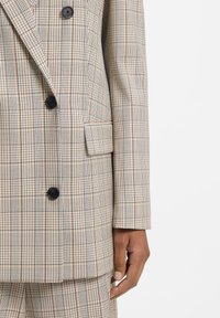 Dobbeltradet blazer i beige ternet mønster med sorte og røde accenter, med to sorte knapper og en sidelomme. Glat stoftstruktur.