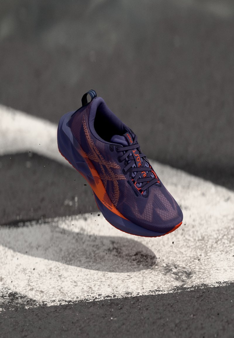 Zapato deportivo morado con acentos naranjas, parte superior de malla, diseño texturizado y una forma delgada y aerodinámica. Cuenta con una lengüeta en el talón.