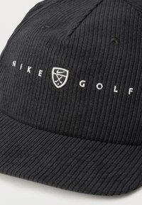 Μαύρο καπέλο από ριμπ ύφασμα με λευκό κεντημένο κείμενο "NIKE GOLF" και μικρό λογότυπο ασπίδας με σήμα swoosh και σταυρωτά μπαστούνια γκολφ.