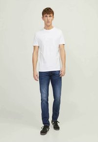 Jovem em pé, vestindo uma t-shirt branca simples, jeans azuis e tênis pretos com riscas brancas, sobre um fundo cinza claro.