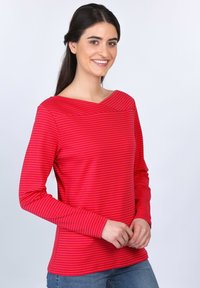 Rotes Langarmshirt mit horizontalen Streifen, das über einen weiten Ausschnitt und eine glatte Textur verfügt. Das Modell trägt es zusammen mit blauen Jeans.