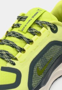 Neon gul atletiksko med mesh-overdel, mørkegrønne snørebånd og det karakteristiske Nike-logo. Har en tekstureret overflade og øje-design.