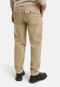 Pantalones de algodón beige con corte recto, con bolsillos traseros con cierres de botón y una pequeña etiqueta de marca en la cintura. Zapatos negros visibles.