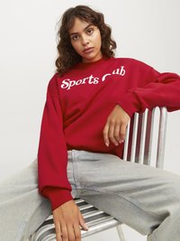 Felpa rossa con la scritta "Sports Club" in bianco, caratterizzata da una vestibilità comoda e polsini a coste, abbinata a jeans di denim blu chiaro.