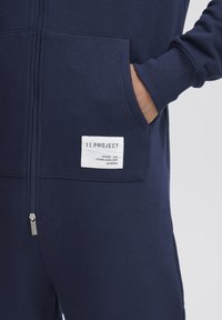 Monosuit azul marino con cremallera, fabricado en un tejido texturizado, con puños acanalados y grandes bolsillos laterales. Presenta una etiqueta de la marca en color blanco en el bolsillo izquierdo.