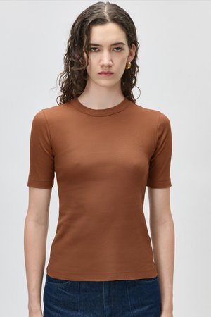 Jeune femme aux cheveux bouclés portant des boucles d'oreilles dorées en forme de créoles, un t-shirt marron ajusté à manches courtes et un pantalon bleu foncé, debout devant un fond uni.