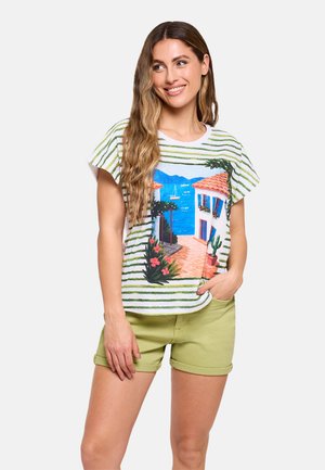 Mujer con el pelo largo que lleva una camiseta de rayas con un estampado de un pueblo costero mediterráneo y pantalones cortos de color verde claro remangados, sonriendo y posando.