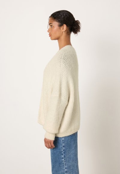 Pull tricoté surdimensionné de couleur crème avec un col en V et des épaules tombantes, associé à un jean en denim bleu clair, vu de profil.