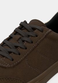 Brown suede sneaker featuring dark gray laces, contrasting stitching, smooth texture, and a minimalist design with a rubber sole.