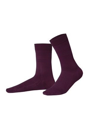 Paire de chaussettes hautes côtelées violet foncé présentée sur fond blanc, l’une légèrement inclinée vers l’avant montrant le pied et la cheville.