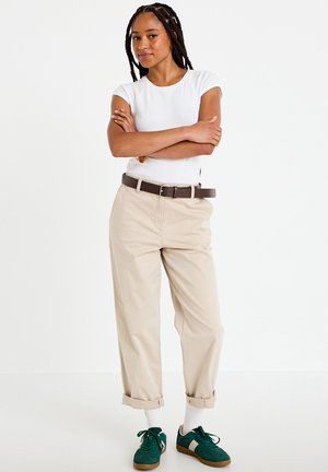 Młoda kobieta z zaplecionymi włosami stoi ze skrzyżowanymi ramionami, ubrana w białą koszulkę, beżowe podwinięte spodnie, brązowy pasek, białe skarpetki i zielone trampki.