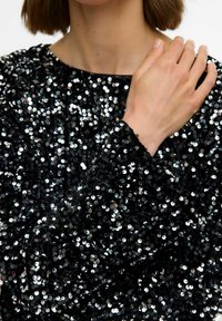 Oseba, oblečena v črno dolgotrajno majico, posuto s sijajnimi črnimi in srebrnimi bleščicami, z eno roko naslonjeno na nasprotno ramo.