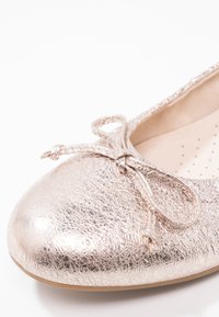 Ballerine plate dorée métallique avec surface texturée et nœud décoratif sur l’orteil arrondi, avec une semelle intérieure légèrement rembourrée.